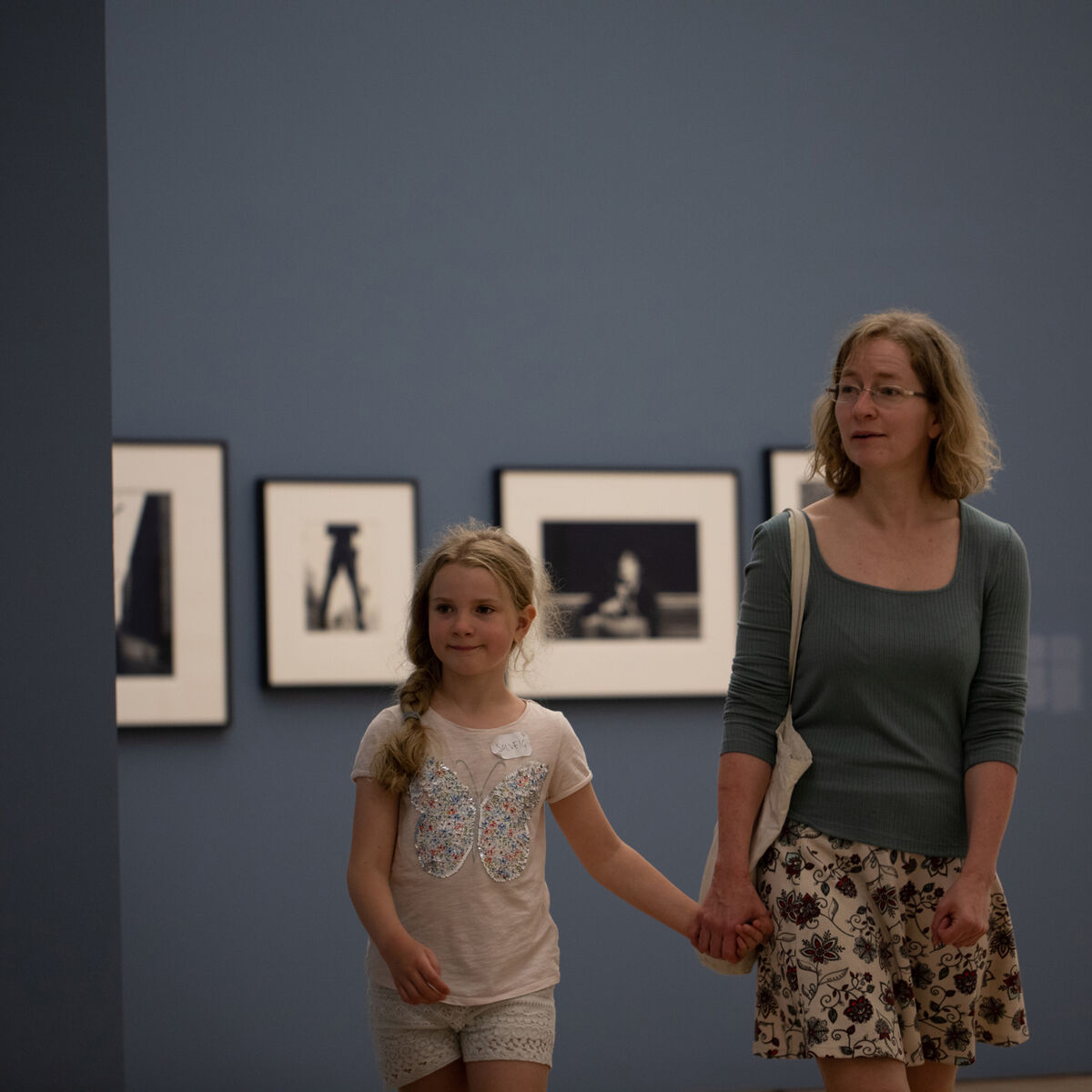 Kvinne og jente hånd i hånd i utstillingslokale, med innrammede fotografier i bakgrunnen